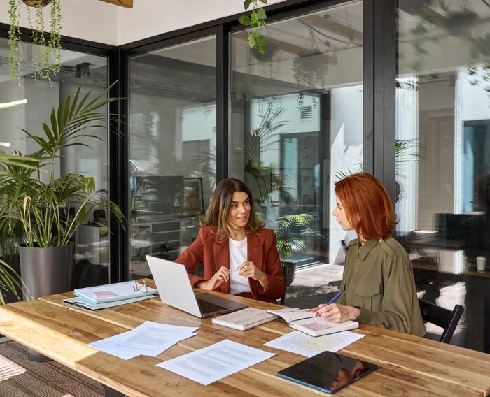 two women sitting at a desk in an office with laptop computers