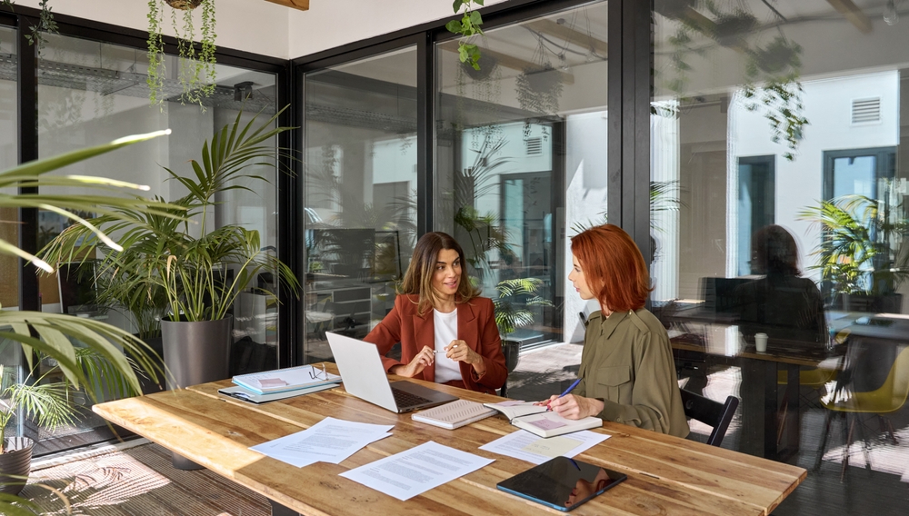 two women sitting at a desk in an office with laptop computers