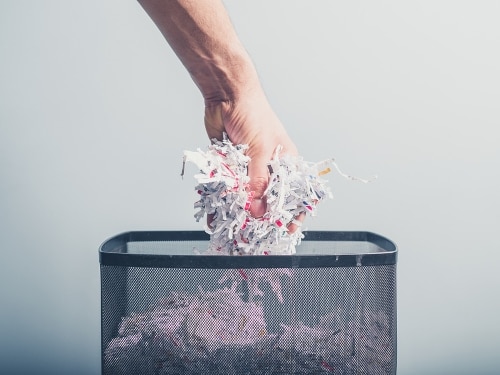 A hand is putting a bunch of shredded paper in a waste paper basket