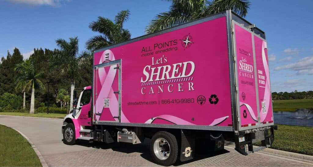 A photo of the All Points Mobile Shredding Pink Truck for a Purpose parked on a paved driveway in front of palm trees and a small fountain in a pond