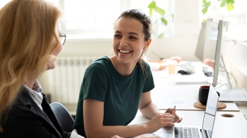 Two women colleagues employees of diverse age sharing points of view at workplace in office.