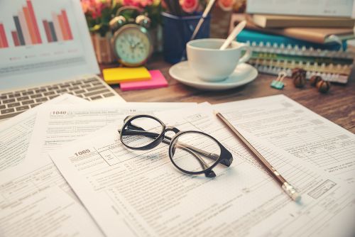 Tax paperwork, glasses, and a laptop are sitting on a desk with a cup of coffee.